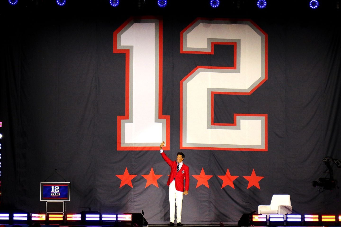 Tom Brady waves under his retired number 12 on stage as he wears his red jacket.