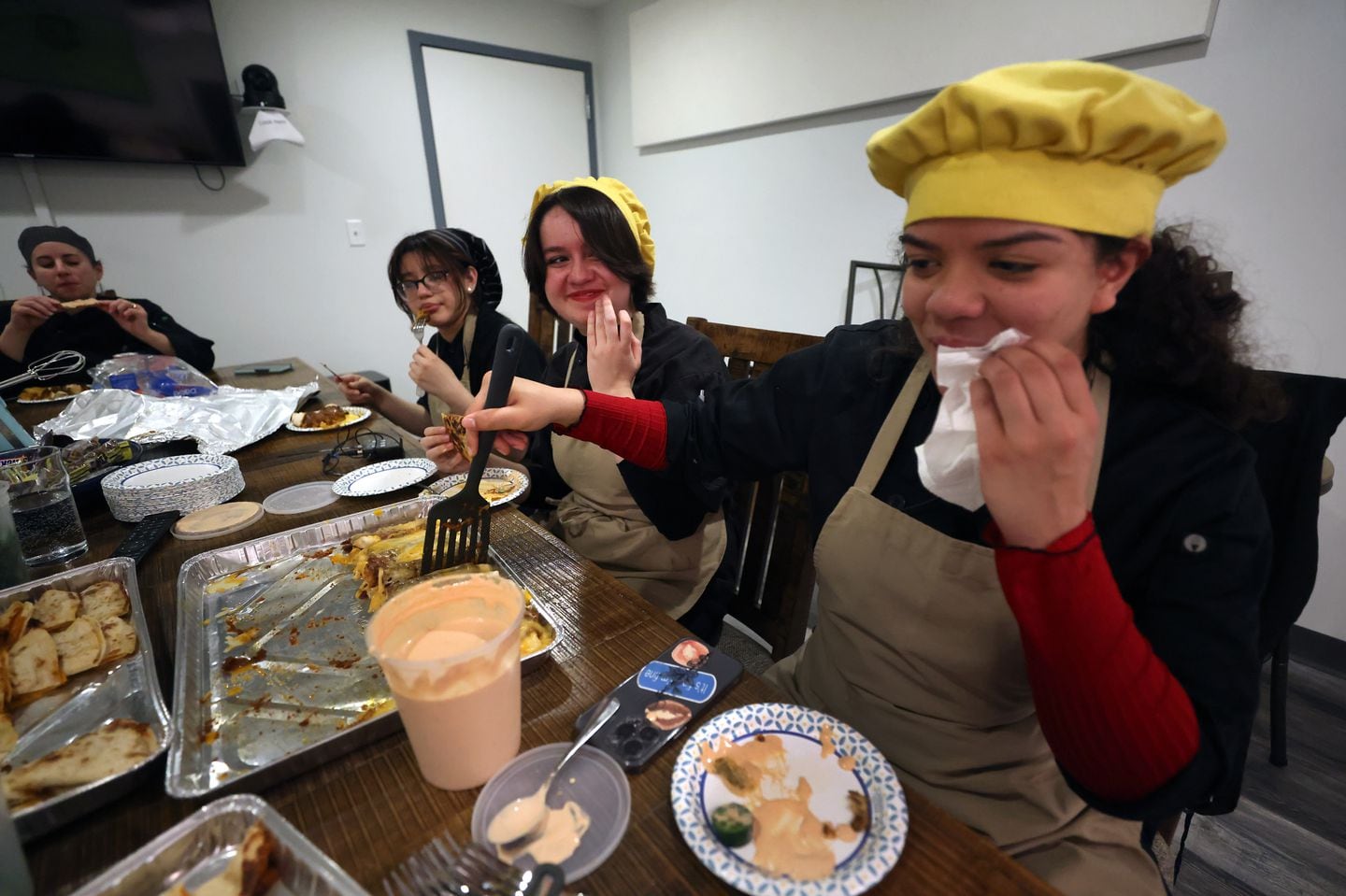 Alexandra Mencia Moradel, uses a napkin to wipe her lip while eating with other students during lunch in the studio where they were cooking for a monthly TV show.  In background is Chef Liz Henderson with Northeast Metro Tech School in Wakefield. Revere teens who are enrolled in the culinary arts program at Northeast Metro Tech School in Wakefield filming the second episode of "Northeast Cooks," a monthly program filmed at RevereTV and shown on local cable.  Each episode of the student-led program spotlights a new dish and features the full scope of culinary arts from knife techniques to mastery in presentation.