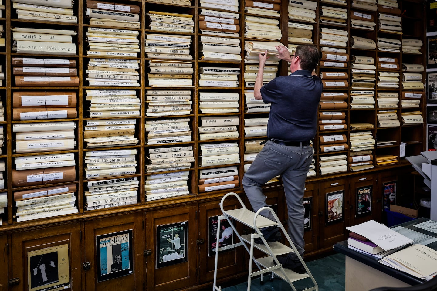 Principal librarian Wilson Ochoa reaches for scores stored in floor-to-ceiling wooden shelving at the Boston Symphony Library, where hundreds of orchestral works by composers like Mahler and Mozart are archived in white folios.