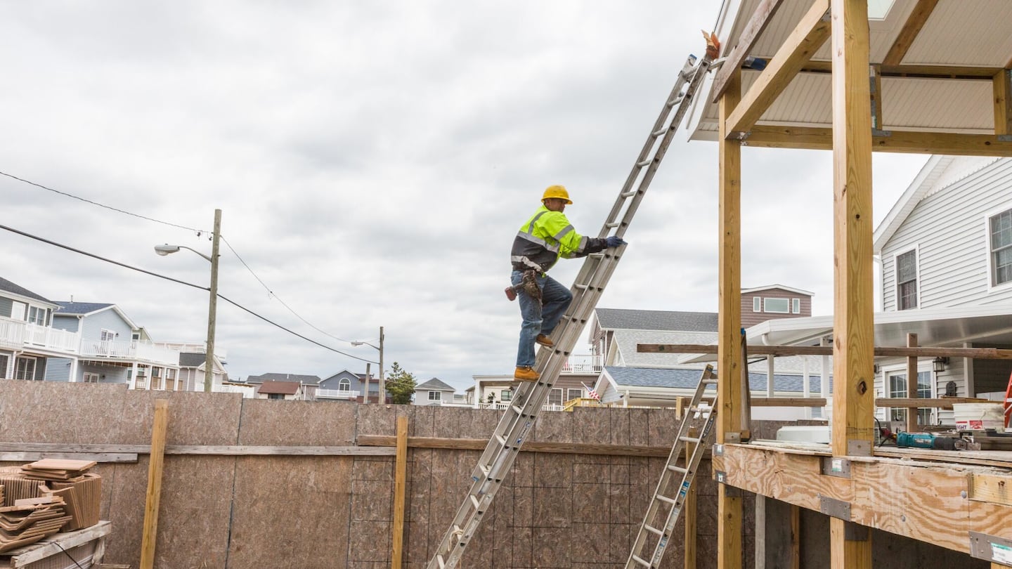 A worker climbs a ladder to install a gutter at the construction site of a house.