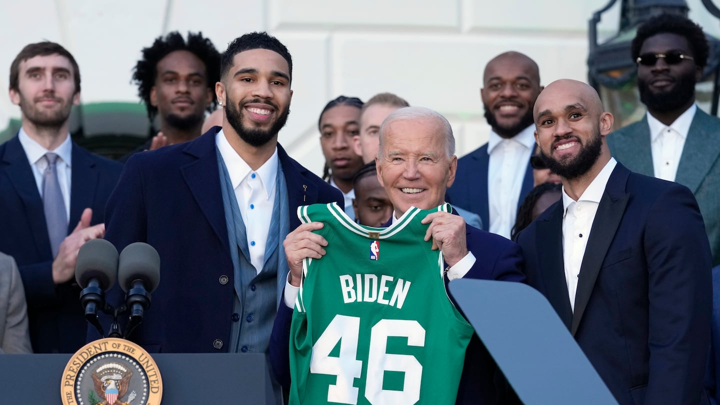 President Joe Biden (center) is flanked by Celtics players Jayson Tatum (left) and Derrick White (right) as he holds up a jersey the team presented to him during an event to celebrate the team's victory in the 2024 NBA Championship on the South Lawn of the White House.