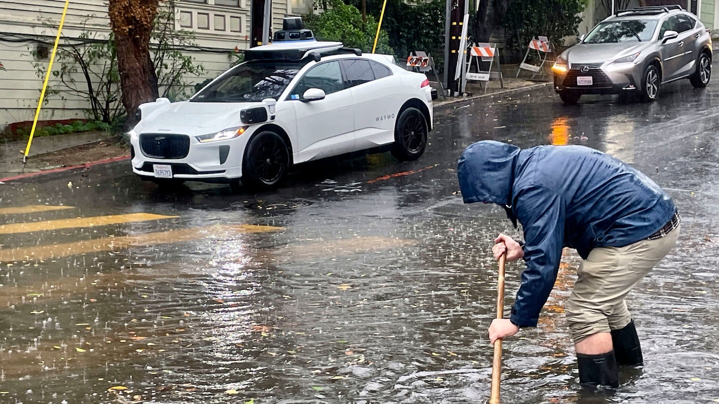 A Mission District resident attempted to unclog a few storm drains on a flooded street as a driverless Waymo taxi passed in the background on Friday in San Francisco.