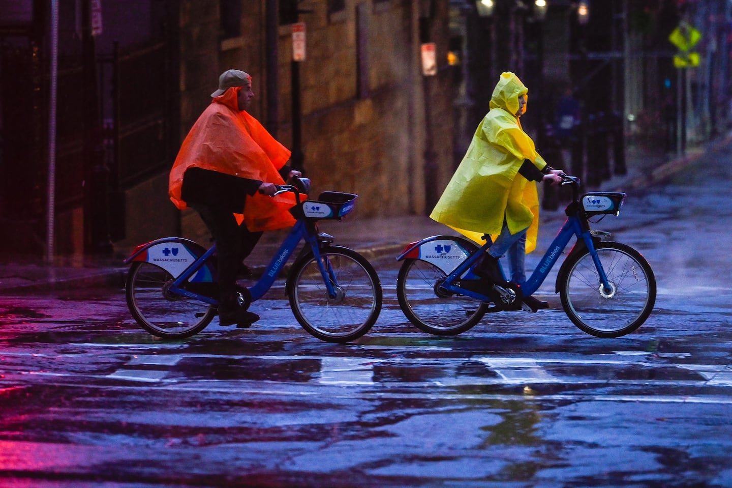 Two people wearing rain ponchos ride BlueBikes down Tremont Street in downtown Boston on a rainy Thanksgiving.